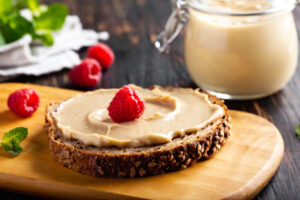 Fetta di pane multicereali con crema spalmabile di anacardi e un lampone su un tagliere di legno, intorno altri lamponi e sullo sfondo un barattolo di crema di anacardi; concept: creme di frutta secca, burro di anacardi