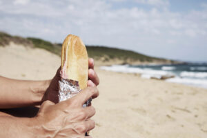 Una persona tiene tra le mani un panino fatto in casa avvolto in alluminio su una spiaggia