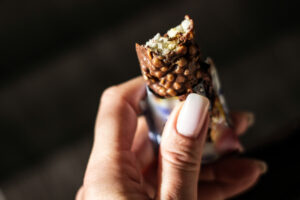 Mano femminile regge una barretta di cereali soffiati ricoperta di cioccolato mezza scartata e mangiata; concept: snack