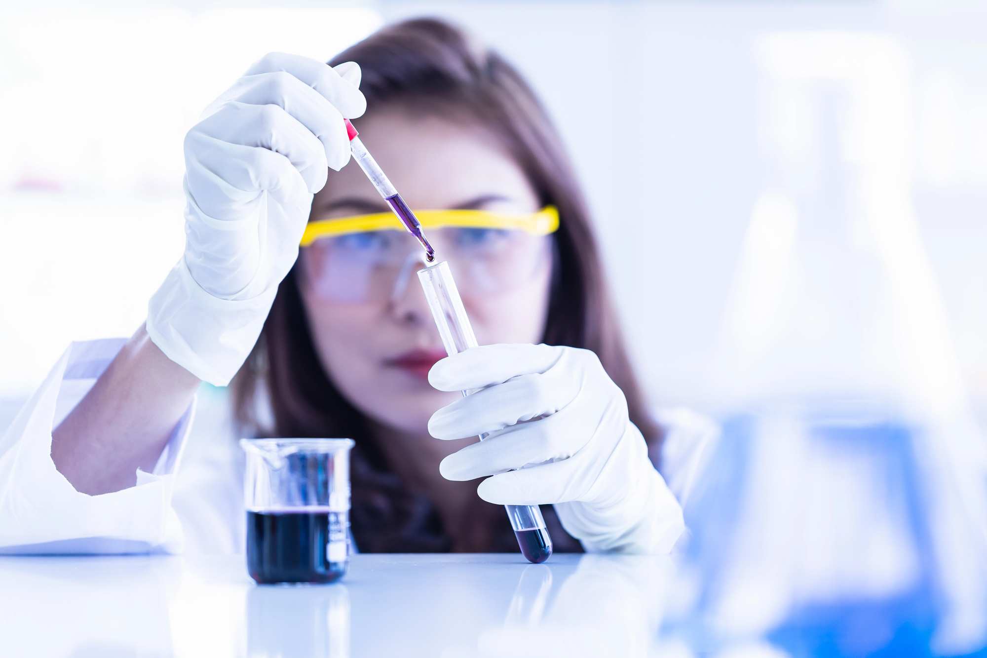 Scienziata o tecnica di laboratorio preleva da una provetta del liquido scuro con una pipetta, sul bancone un becher con altro liquido scuro; concept: cacao, cioccolato, caffè sostanze chimiche