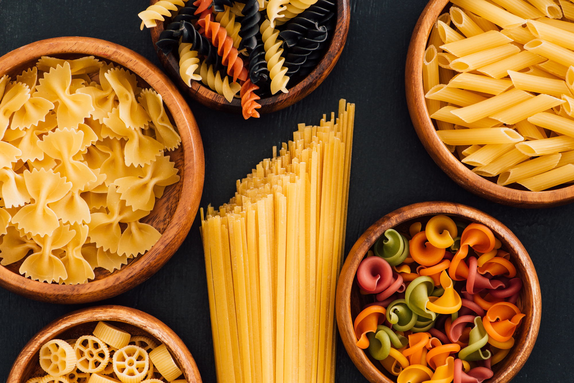 Vari formati di pasta in ciotole di legno su un piano scuro: al centro linguine, intorno ruote, farfalle, fusilli, penne rigate