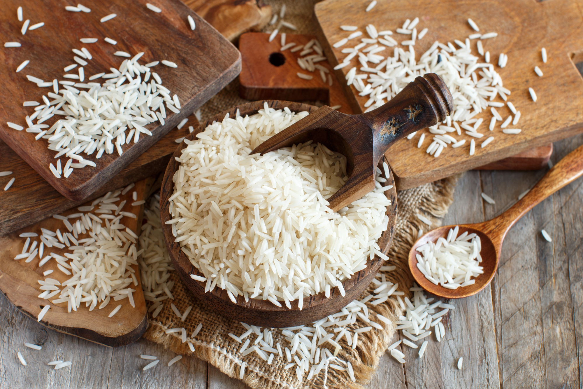 Una ciotola di legno piena di riso basmati con una paletta di legno immersa nel riso; tutto intorno chicchi e mucchietti di riso basmati sparsi su taglieri e cucchiai di legno