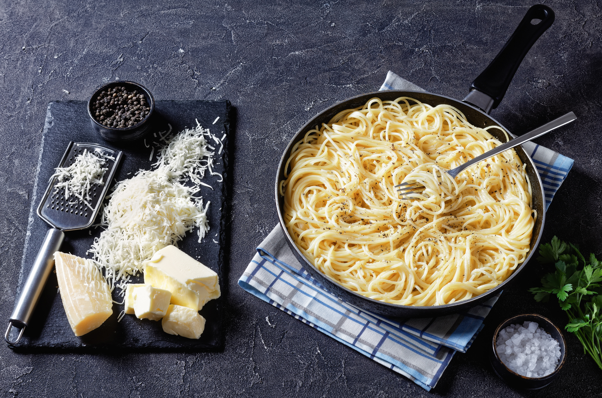 Una padella di spaghetti cacio e pepe con forchetta, appoggiata su uno strofinaccio, accanto a un tagliere con formaggio grattugiato e a pezzi e una ciotola di grani di pepe; concept: pasta