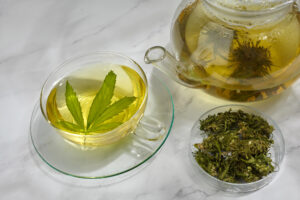 Una tazza di vetro piena di té, infuso o tisana con foglia di canapa, accanto a una ciotola di infiorescenze di cannabis e una teiera di vetro con infiorescenze secche in ifusione