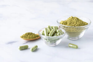 Capsule di integratore di moringa in una ciotolina di vetro, accanto polvere di moringa in una ciotola di vetro e in un cucchiaio di legno