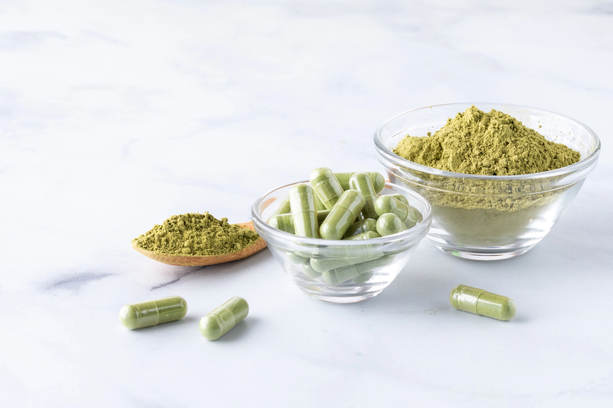 Capsule di integratore di moringa in una ciotolina di vetro, accanto polvere di moringa in una ciotola di vetro e in un cucchiaio di legno