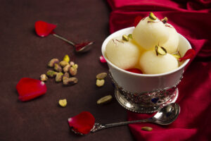 Rasgulla, dolci tipici indiani in una ciotolina d'argento accanto a un cucchiaino, su una tovaglia rossa, sparsi petali di rosa e pistacchi